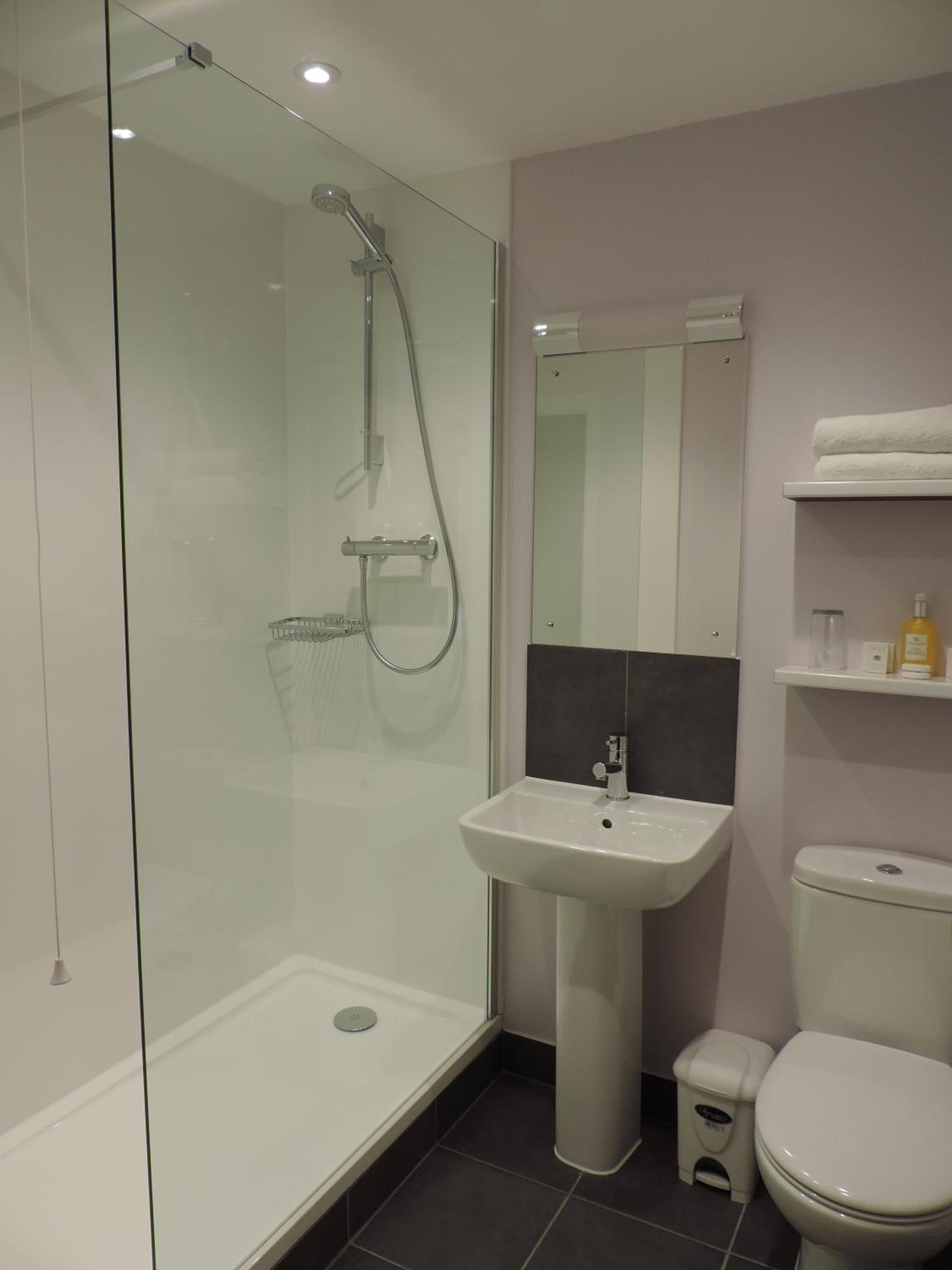 A modern bathroom with a glass-enclosed shower, a pedestal sink, a toilet, and a small trash bin.