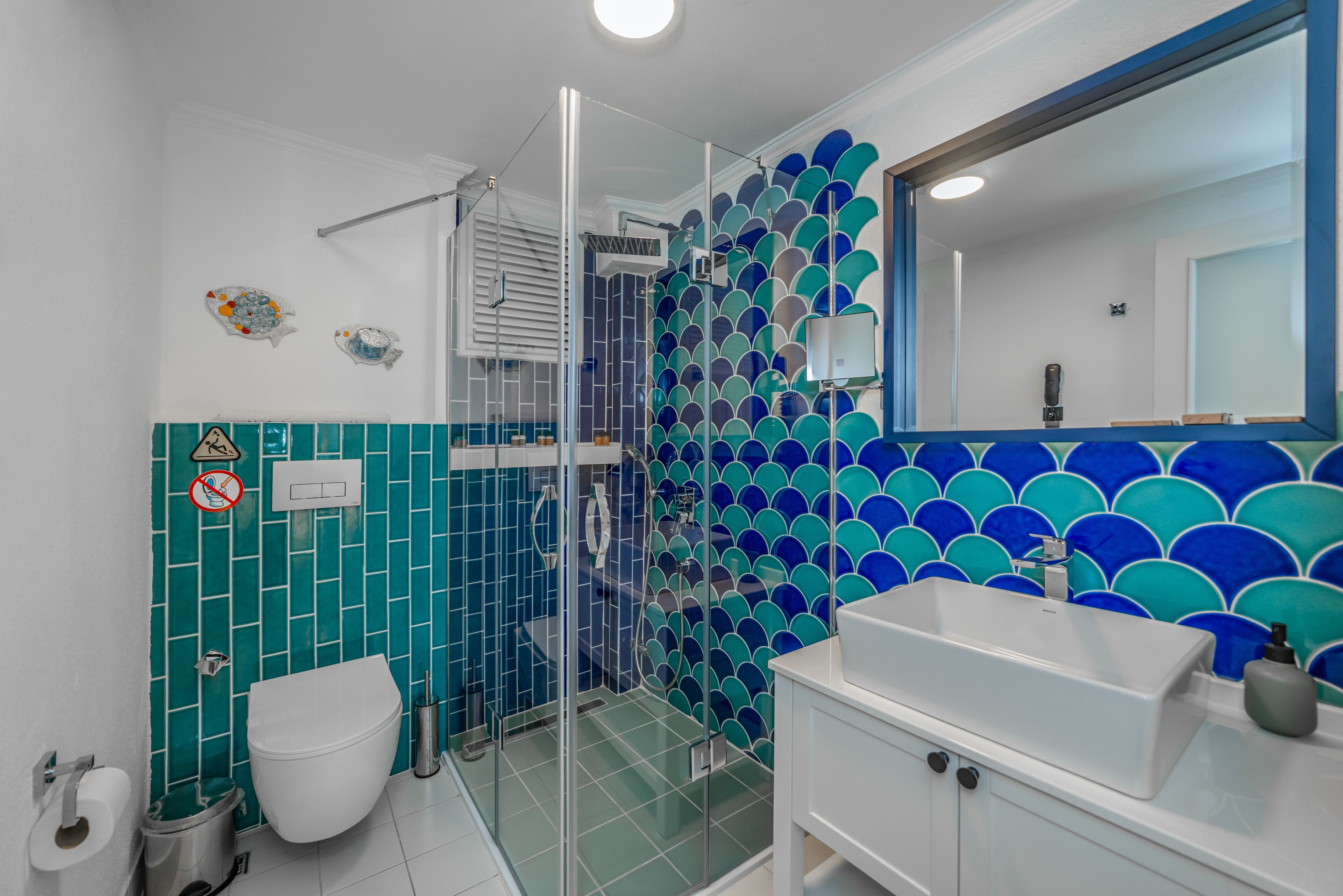 A bathroom with a glass-enclosed shower, a white sink, and a tiled wall with a blue and green pattern.