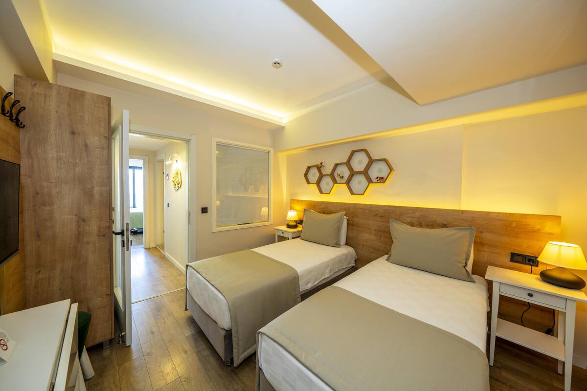 A hotel room with two beds, a wooden headboard, a wall-mounted hexagonal light fixture, and a bedside table with a lamp.