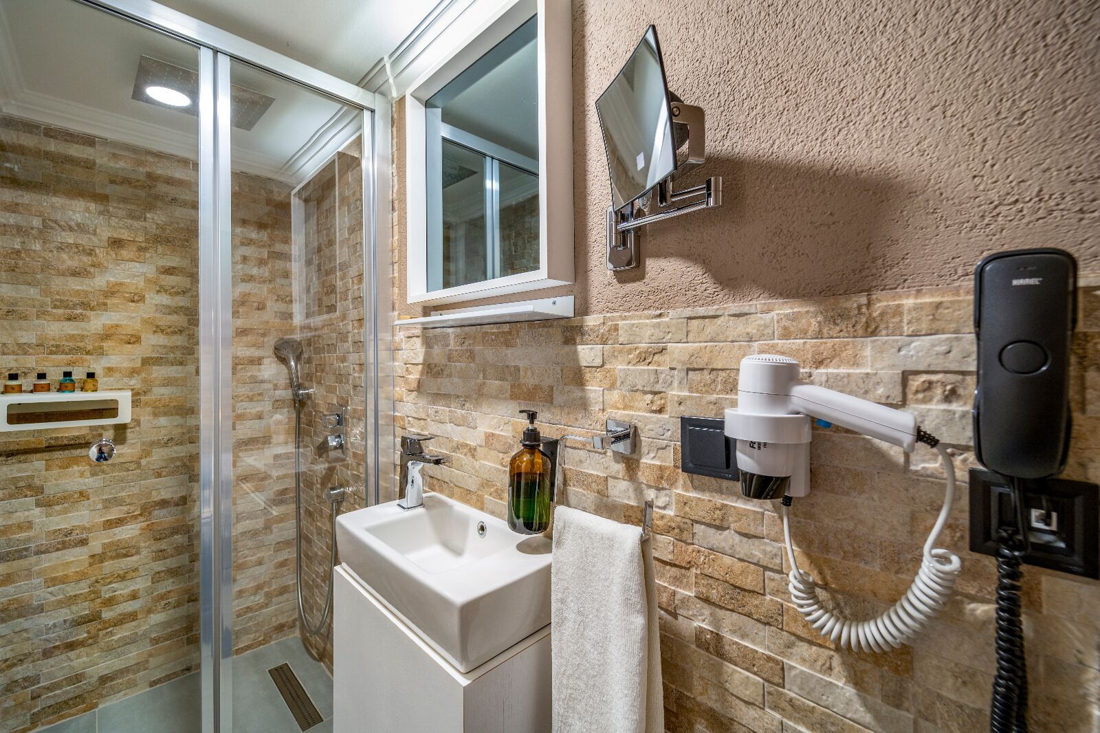 Un baño con cabina de ducha de vidrio, lavamanos con dispensador de jabón, toallero, secador de pelo y teléfono.