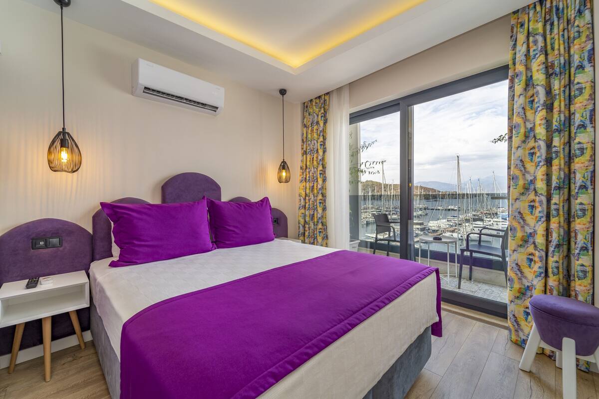 A hotel room with a large bed, purple bedding, a nightstand, a desk, and a view of a marina through a large window.