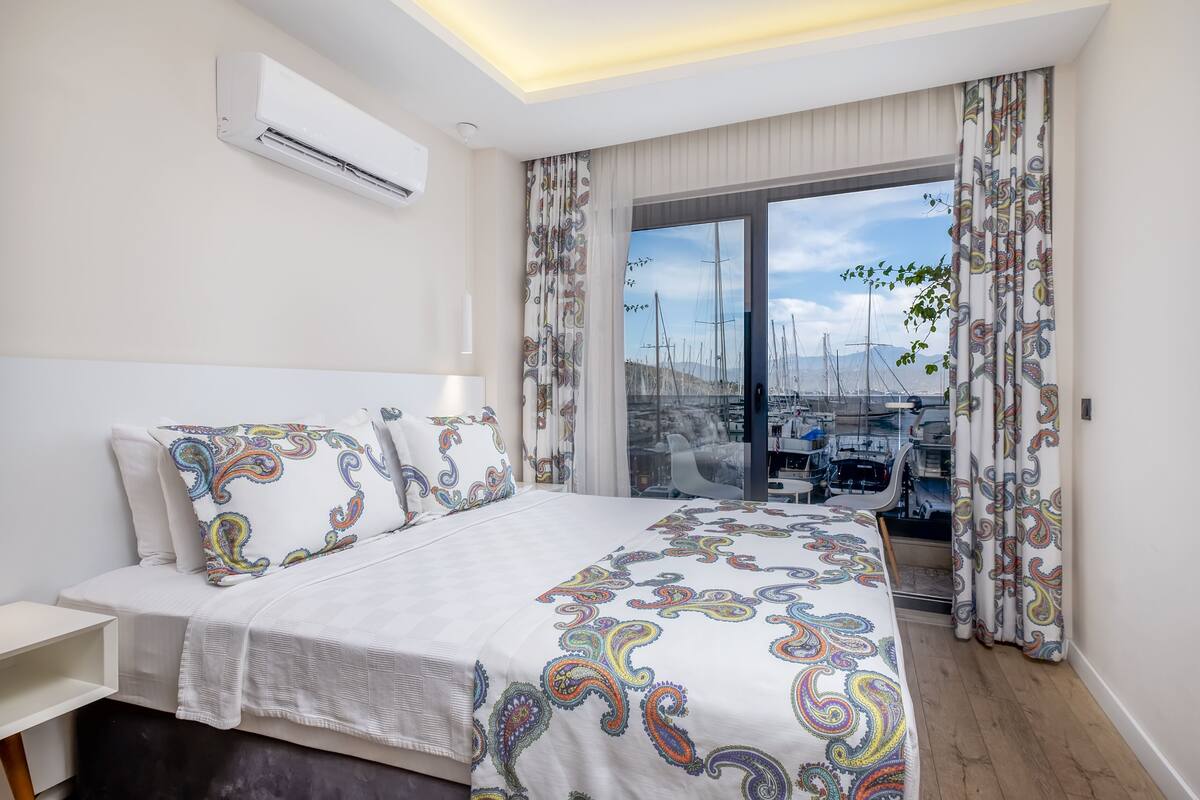 A modern hotel room with a large bed, a view of boats through a glass door, and a ceiling air conditioner.