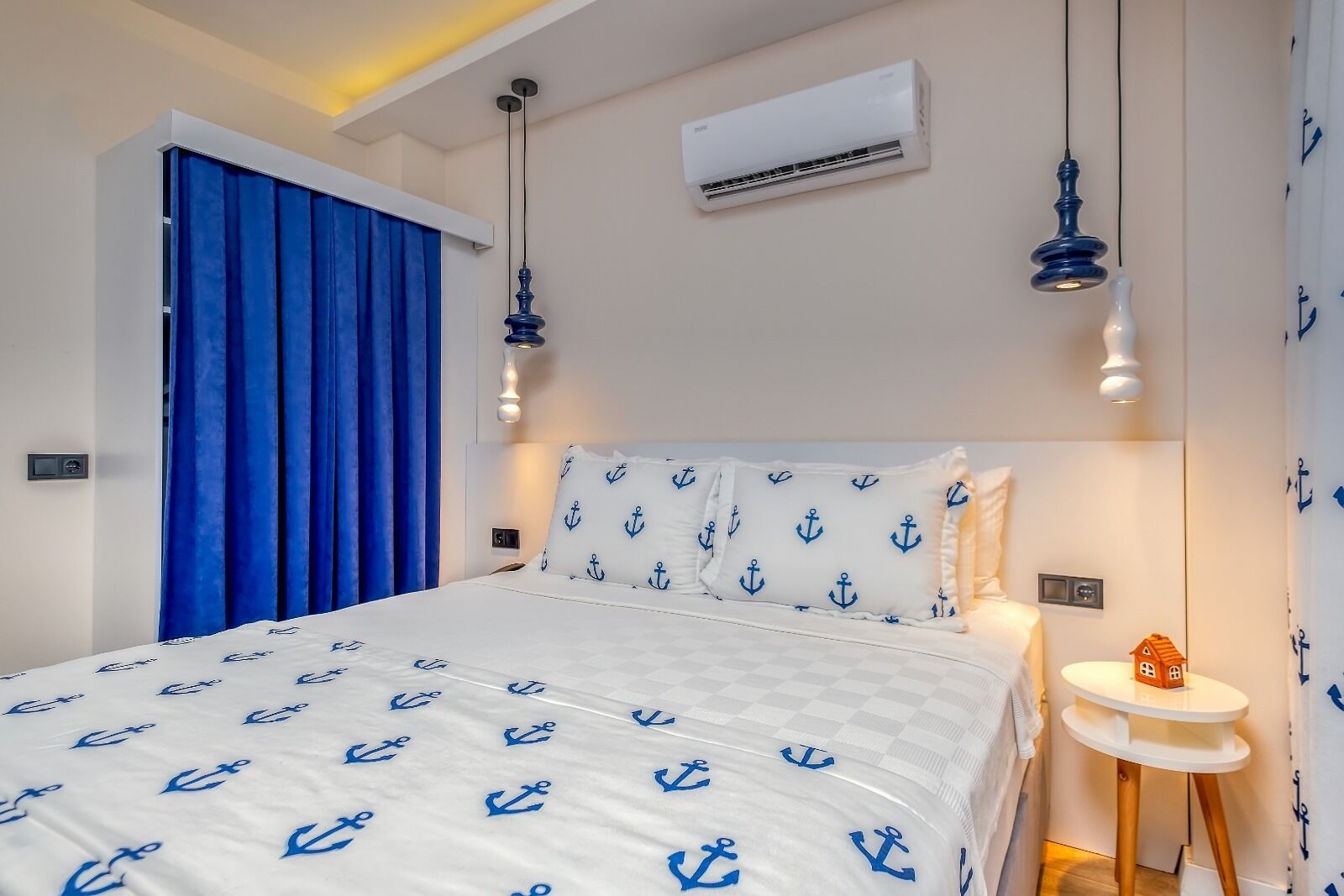 Une chambre à coucher avec un lit, des rideaux bleus, un climatiseur et une petite table sur laquelle est posé un modèle de maison.