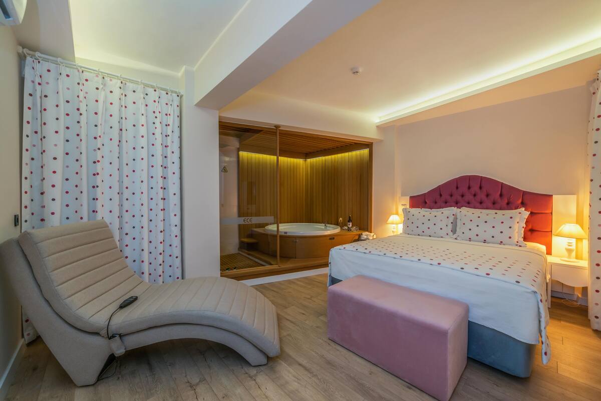 A modern hotel room with a large bed, a sitting area, a bathtub, and a wooden panel wall.