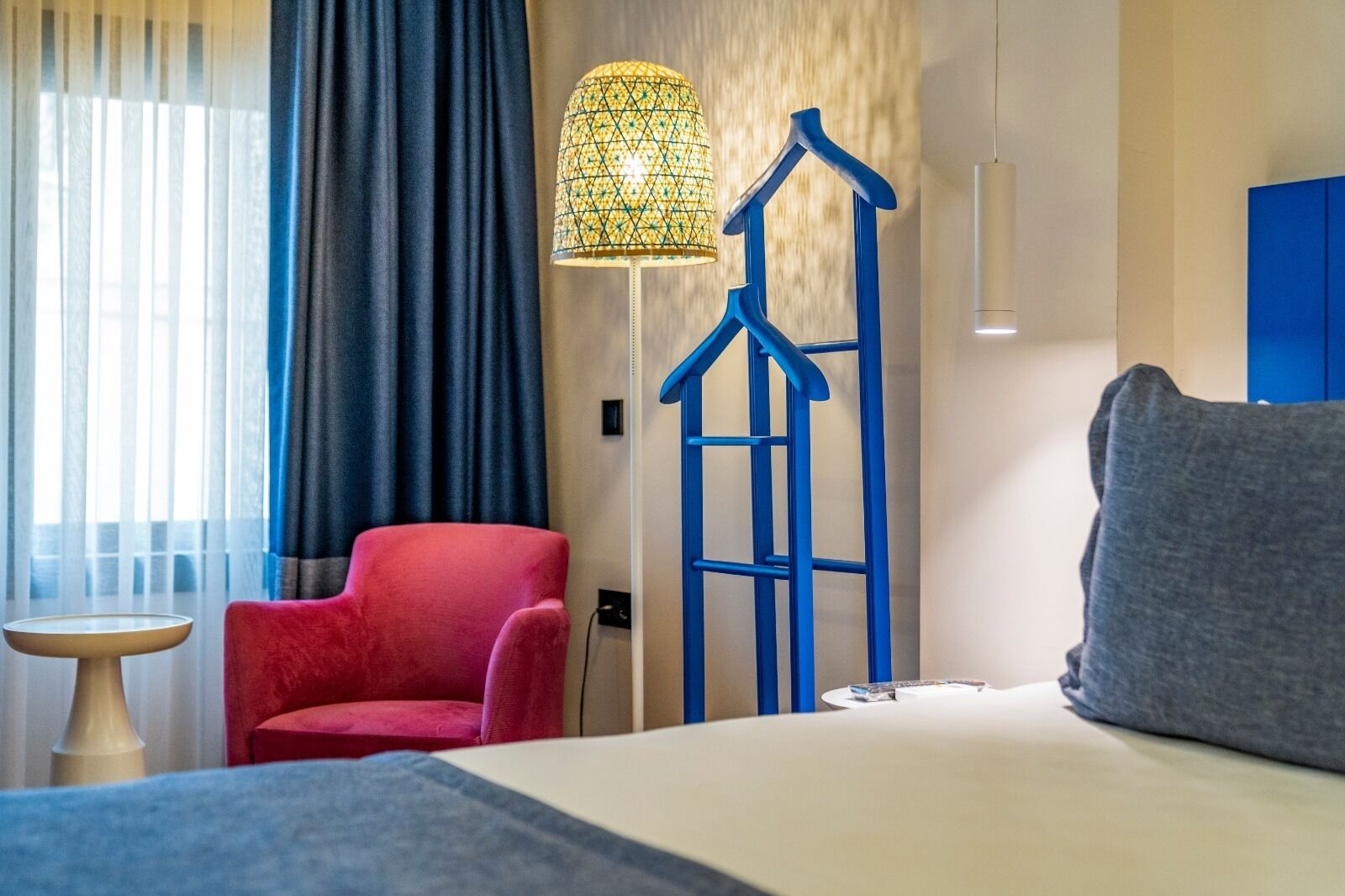 Una habitación de hotel con una cama, un sillón rojo, una silla colgante azul, una mesita y una lámpara.