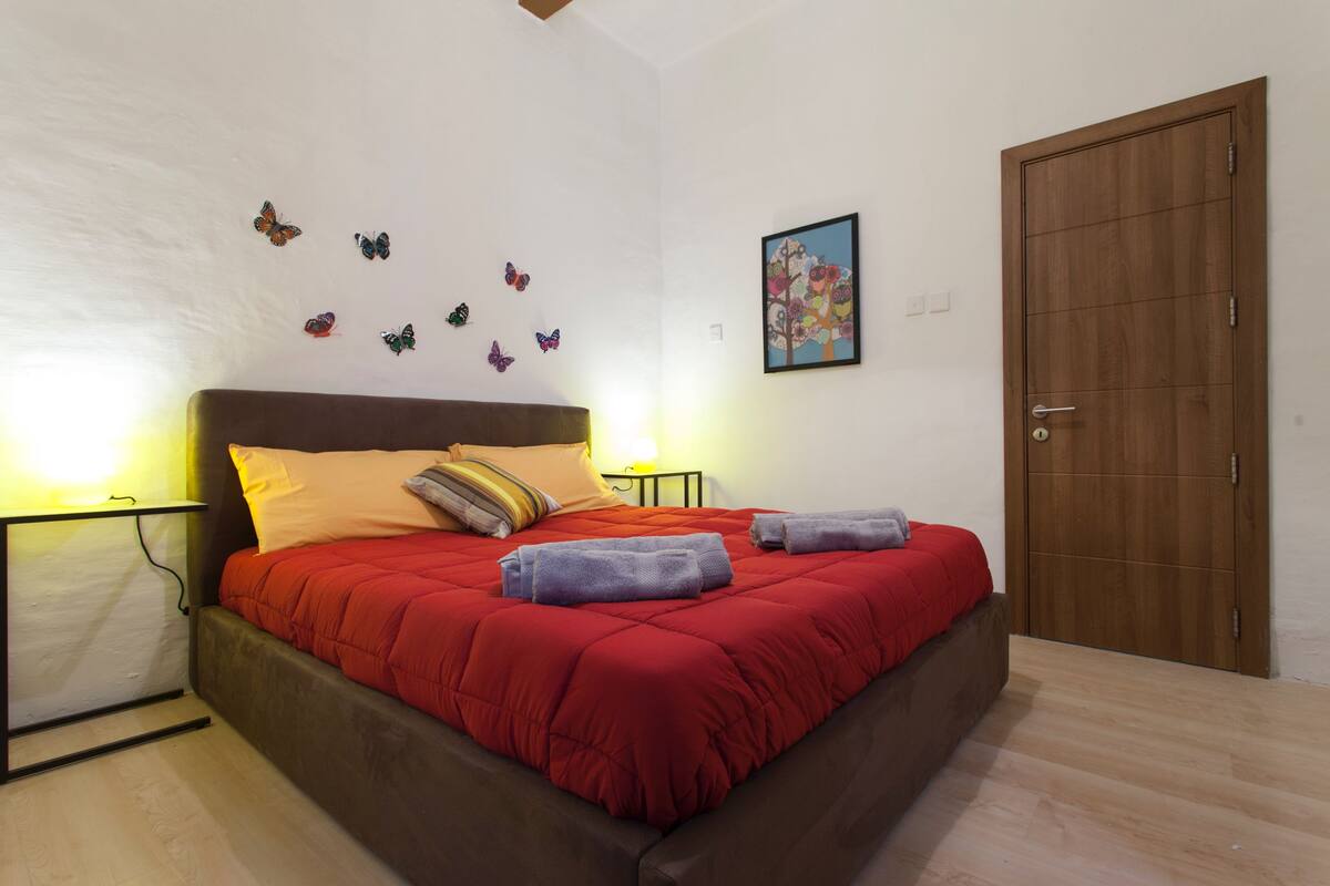 Quarto com colcha vermelha, várias almofadas, porta de madeira e detalhes na parede, incluindo borboletas e uma moldura com fotografia.