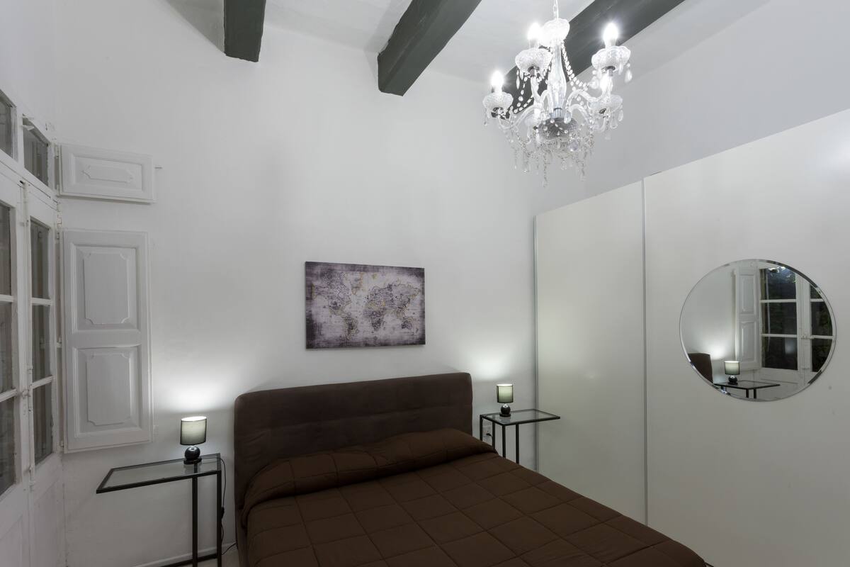 Um quarto com uma cama, um candelabro, um espelho redondo e uma pequena mesa com uma luminária.