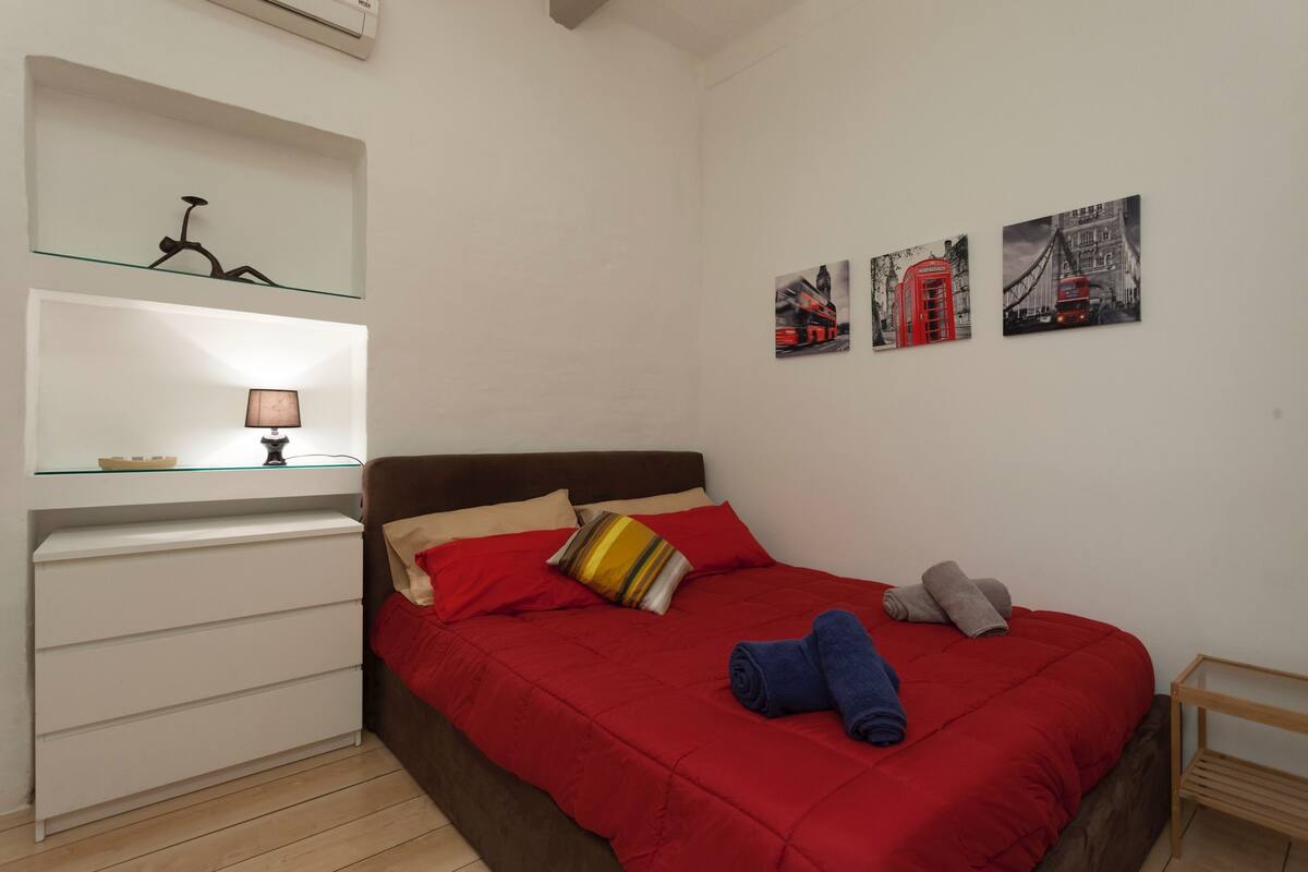Quarto com colcha vermelha, mesa de cabeceira de madeira, prateleira de vidro com abajur e três quadros pendurados na parede.