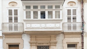 Ein Gebäude mit einer klassischen Fassade, das einen zentralen Eingang mit Doppeltüren, flankiert von Fenstern mit Rollläden, sowie einen Balkon mit aufwendigen Geländern zeigt.