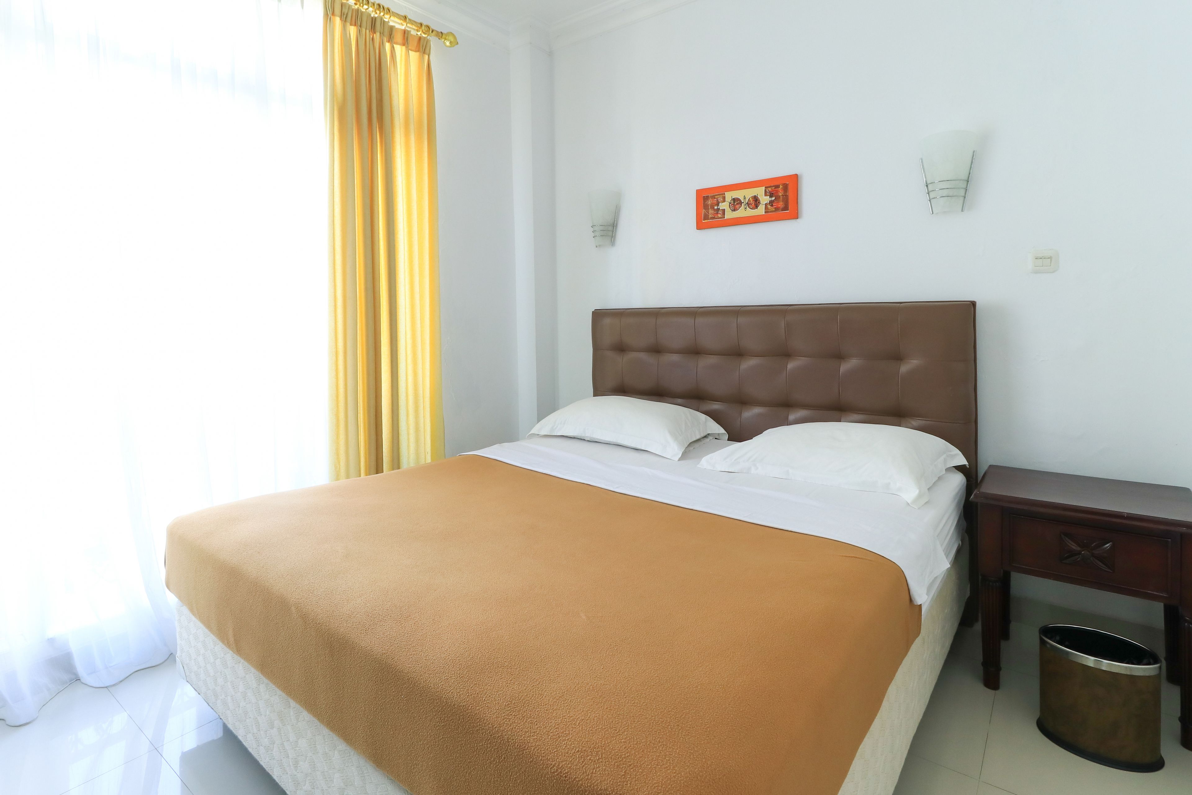 A neatly made bed with a brown bedspread, a wooden nightstand with a trash can, and a wall-mounted light fixture.