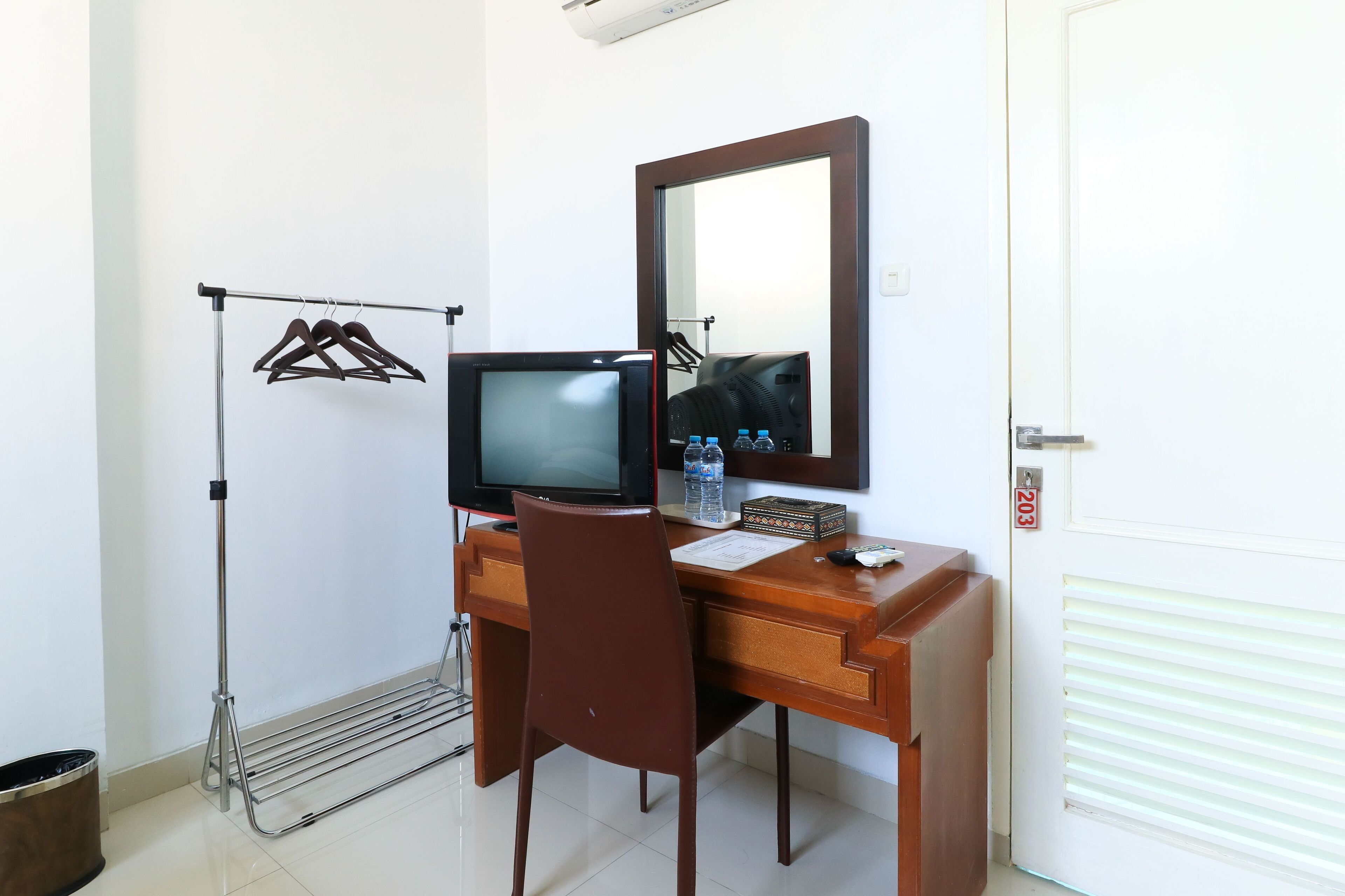 A hotel room with a wooden desk, a television, a mirror, a clothes drying rack, and a door with a number.