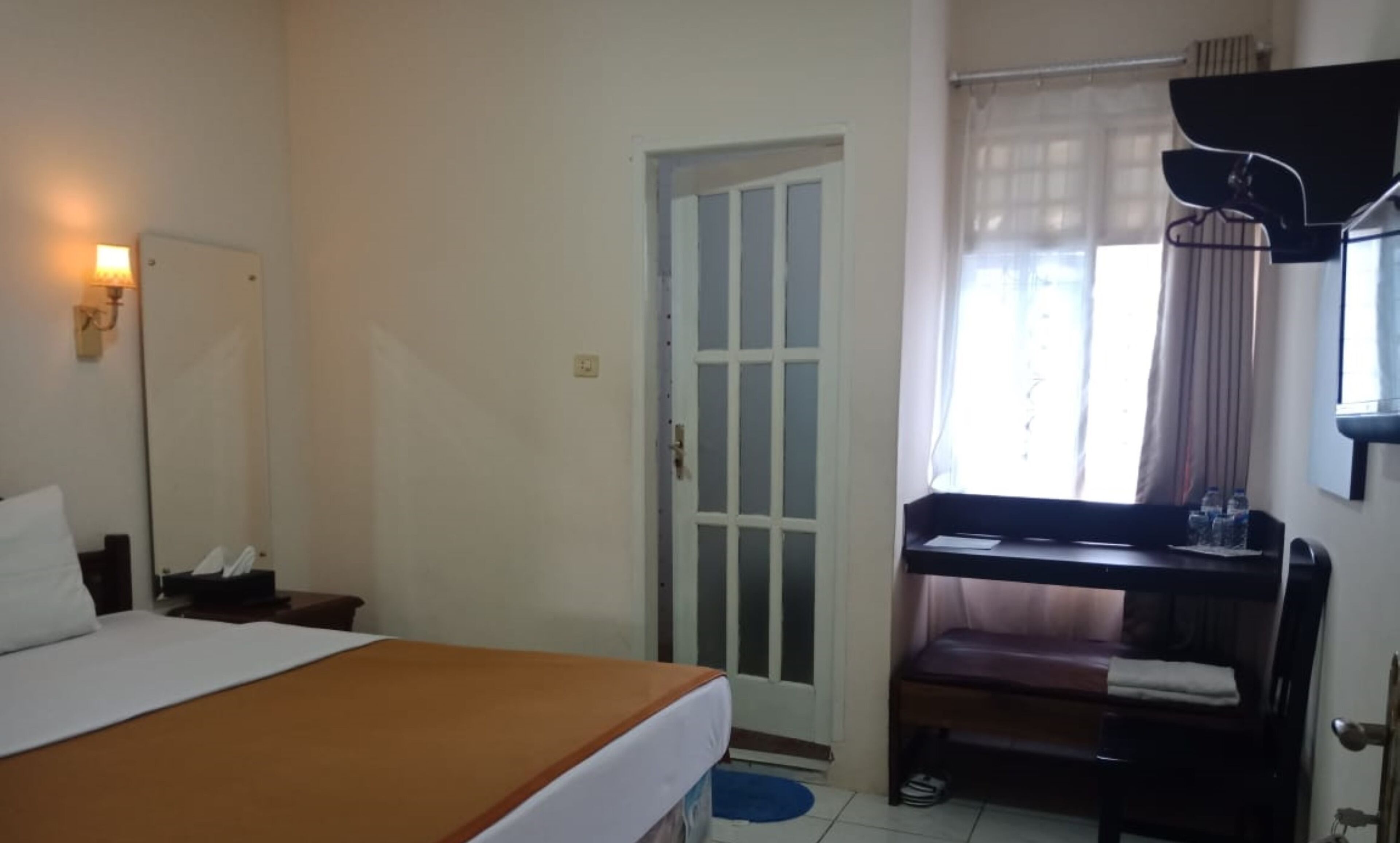 A hotel room with a bed, a desk with a chair, a mirror, a door, and a window with curtains.
