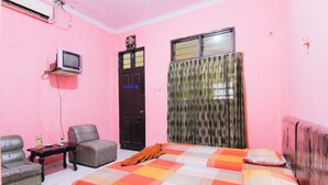 Una habitación con paredes rosadas, una cama, dos sillas, una mesita y un televisor montado en la pared.