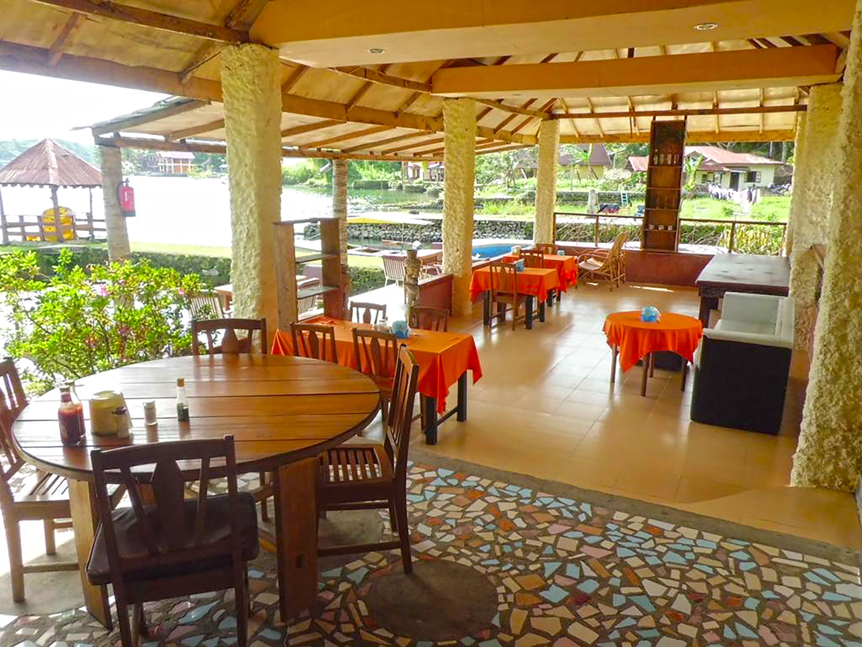 Un restaurante al aire libre con mesas y sillas redondas, cubierto por un techo de madera y un suelo de mosaico.