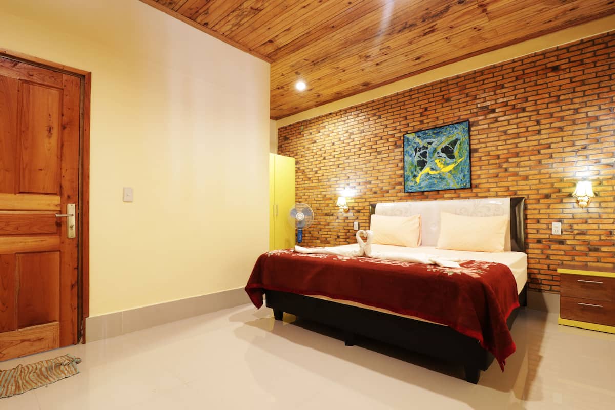 Quarto com teto de madeira, parede de tijolos, cama com roupa de cama branca e manta vermelha, além de um quadro sobre a cama.
