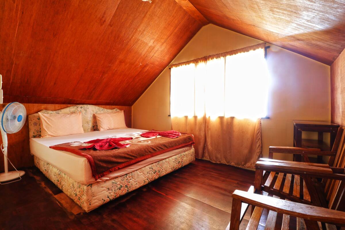 Quarto com teto de madeira, cama com roupa de cama floral, um ventilador de cabeceira e janela com cortinas.