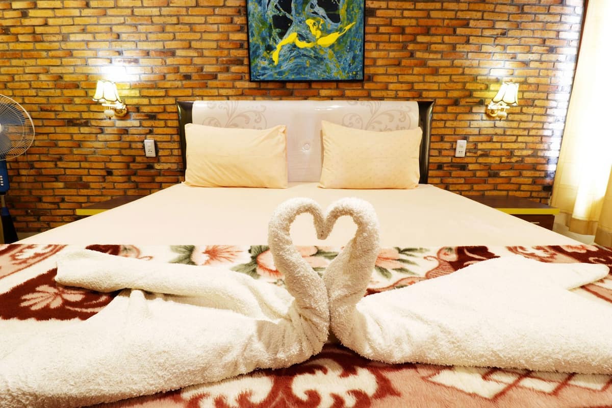Quarto de hotel com parede de tijolos, duas luminárias fixadas na parede, cama com colcha florida e toalhas dispostas em formato de coração.