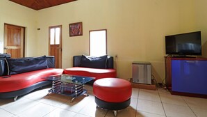Een woonkamer met een rode bank, een glazen salontafel, een televisie op een kast en een kleine koelkast.