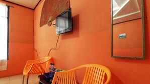 A room with orange walls, two orange chairs, a small table with a bottle, a television mounted on the wall, and a framed picture. - Bangkit Nan Jaya (Berastagi)