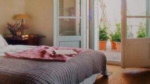 Une chambre à coucher avec un lit, une table de chevet, une lampe et une vue sur un jardin, visible à travers une porte ouverte.