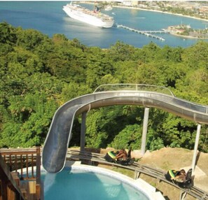 Slide into fun at this resort, where a water slide meets a serene pool surrounded by lush greenery and a stunning ocean view.