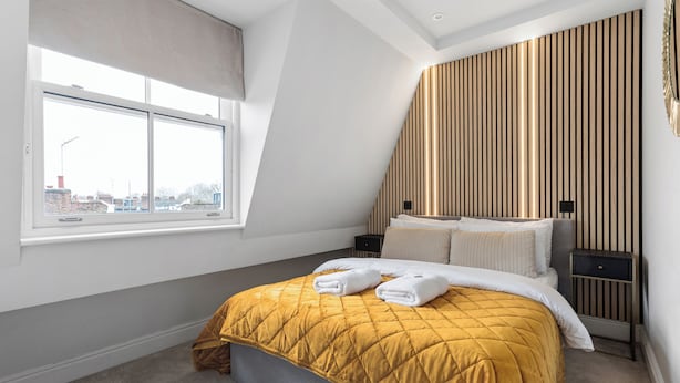 Ein Schlafzimmer mit großem Fenster, einem Bett mit gelber Steppdecke, einem Nachttisch und einer Wand mit vertikalen Holzleisten.