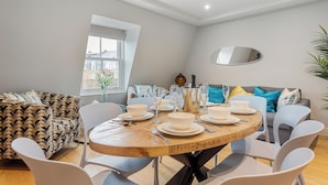 Ein modernes Wohnzimmer mit einem runden Holzesstisch, gedeckt für vier Personen, umgeben von weißen Stühlen. Ein gemustertes Sofa und ein graues Sofa mit blauen und gelben Kissen. Ein großes Fenster bietet einen Ausblick nach draußen.