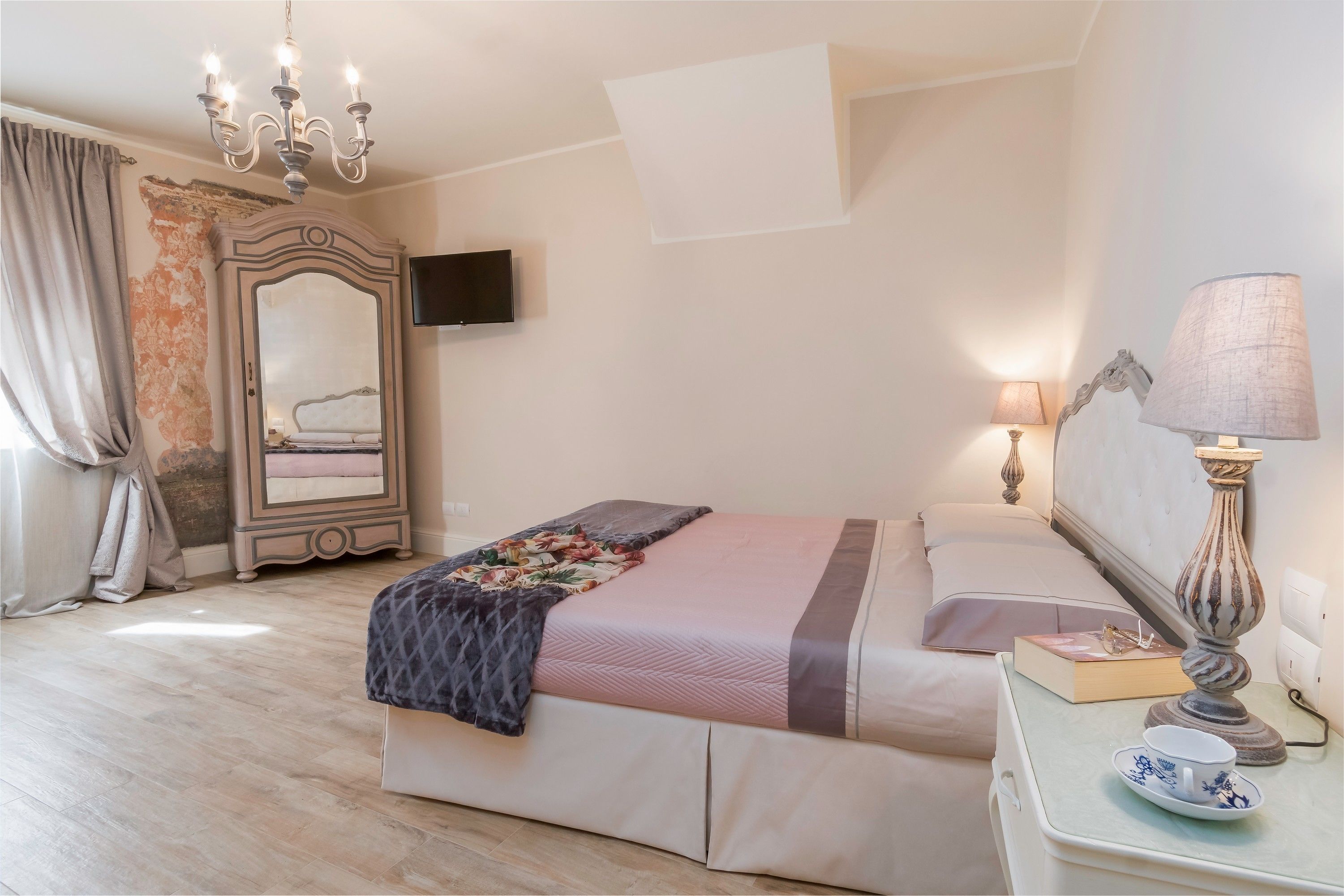 Un dormitorio ordenado con cama, lámparas de noche, una mesita de noche con un libro y una taza, un espejo y un candelabro.