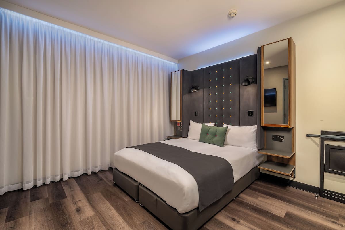 Moderni hotellihuone, jossa on suuri sänky, yöpöytä, peili ja ikkuna, jossa on verhot.