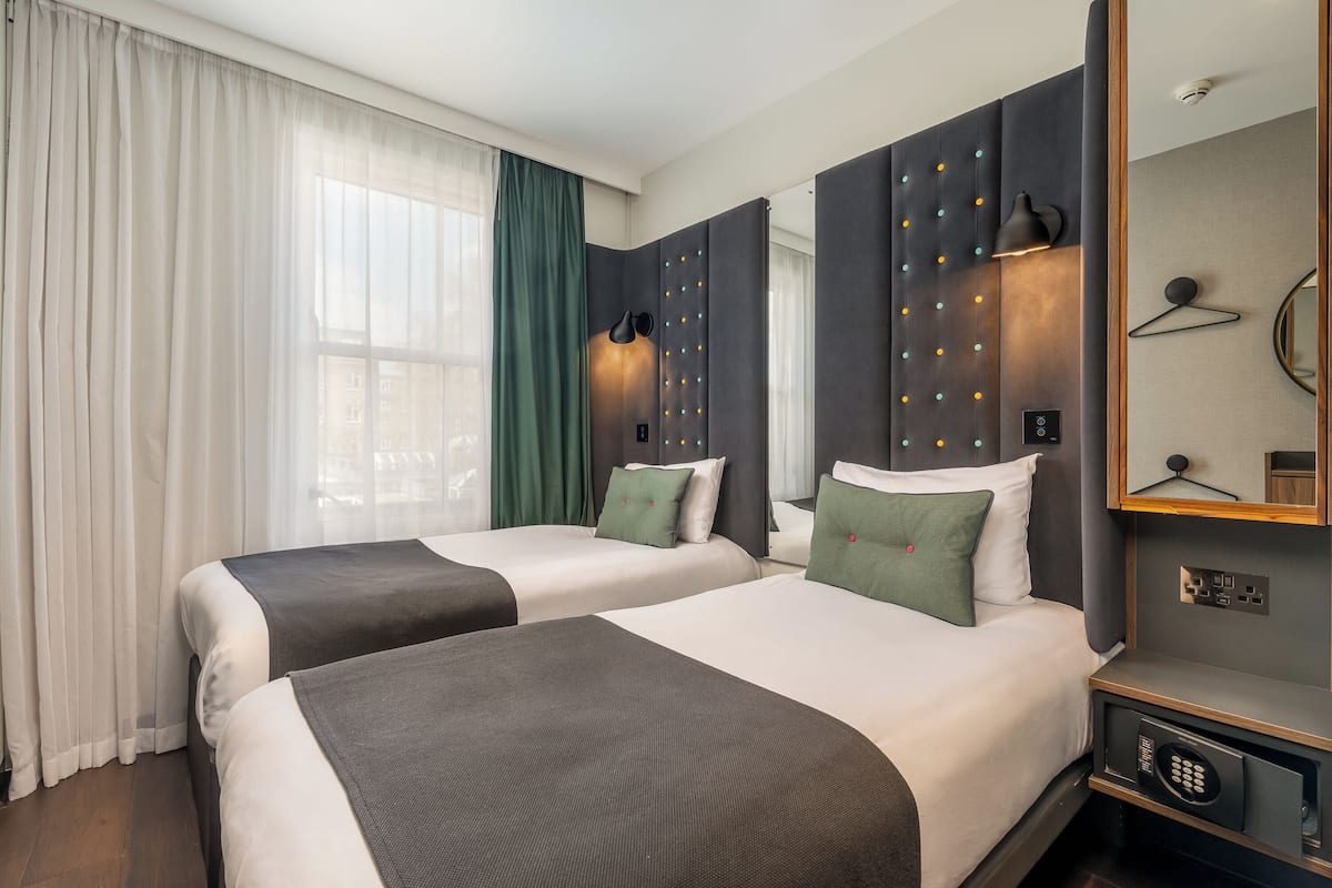 Hotellihuone, jossa on kaksi sänkyä, suuri ikkuna läpikuultavilla verhoilla, seinään kiinnitetty valaisin, peili ja pieni yöpöytä.
