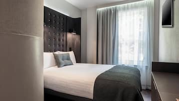 Une chambre d’hôtel moderne avec un grand lit, une tête de lit sombre, un téléviseur à écran plat et une grande fenêtre agrémentée de rideaux légers.