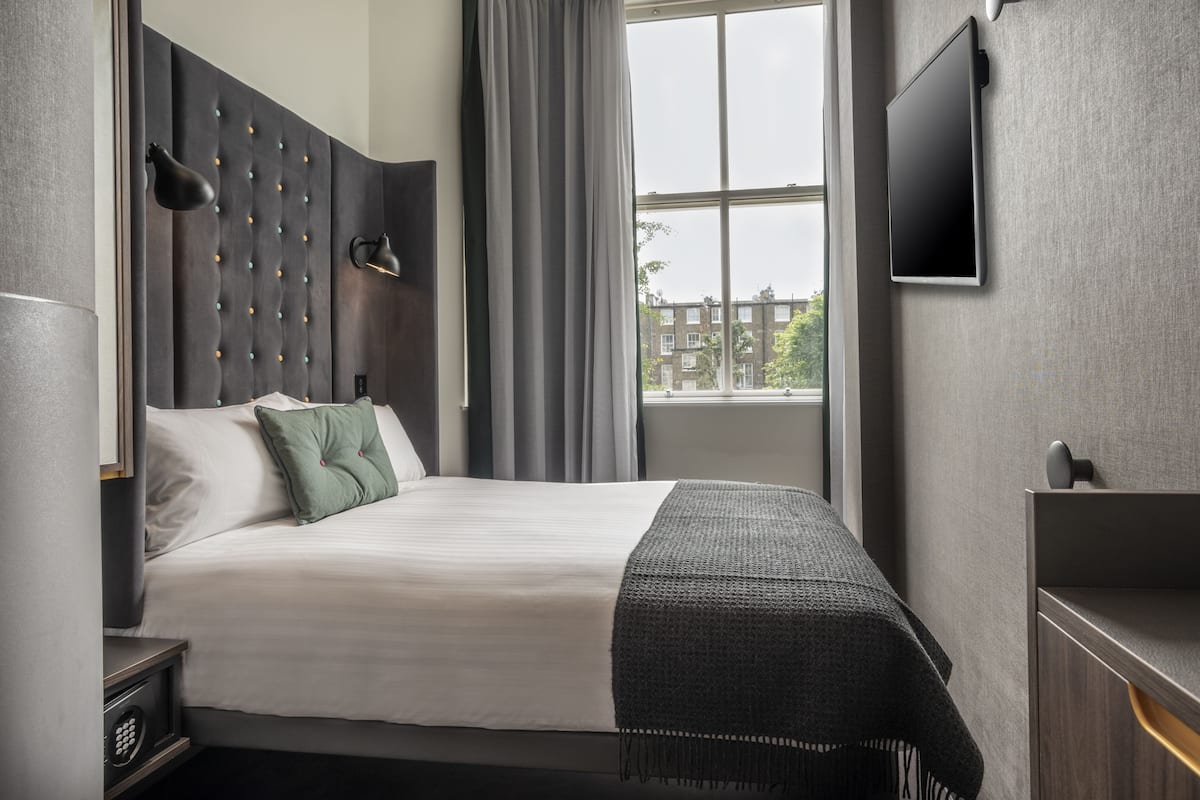 Moderni hotellihuone, jossa on suuri sänky, litteänäyttöinen televisio, ikkuna, josta on näkymä rakennuksiin, ja seinälle asennettu lamppu.