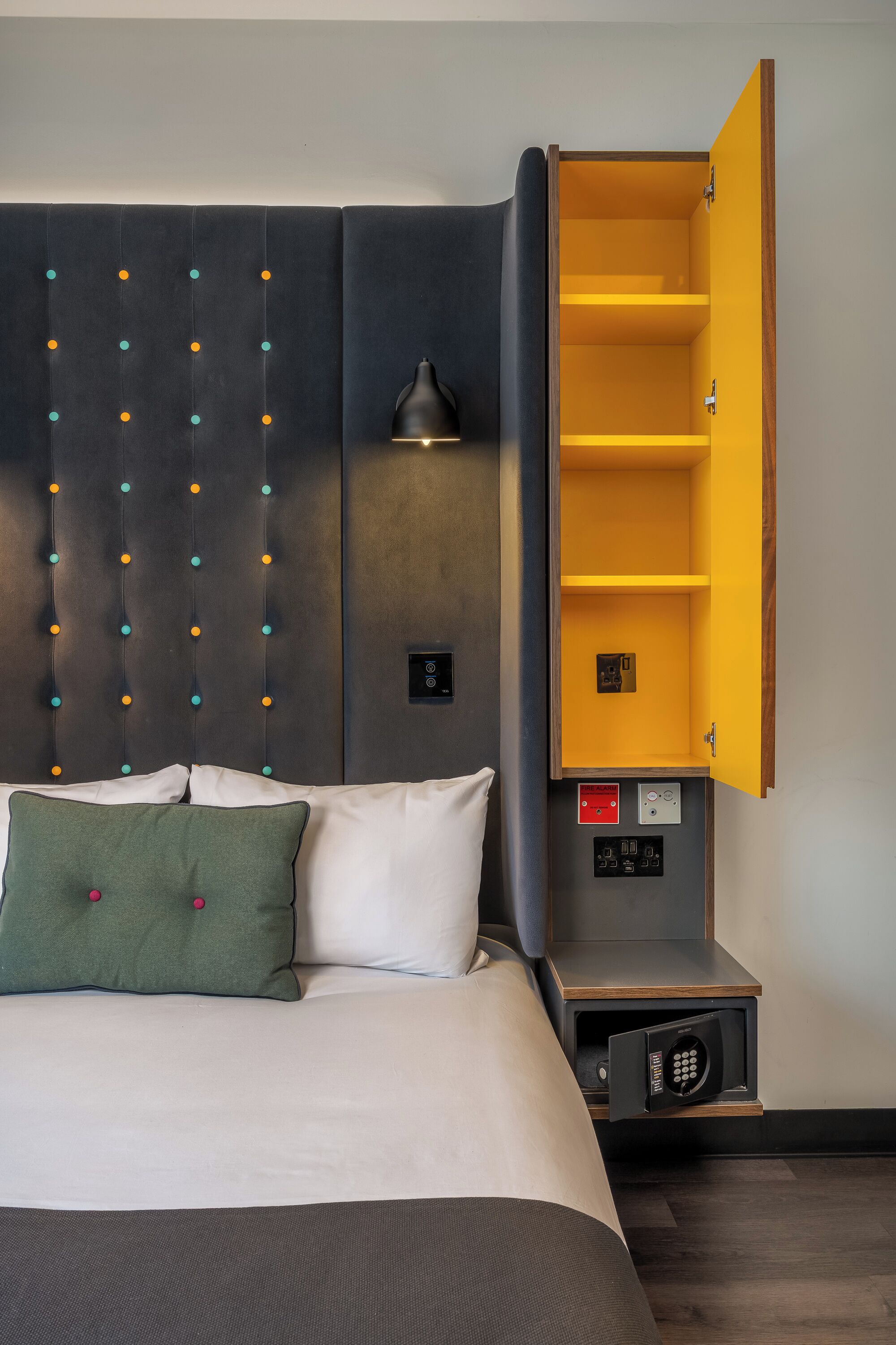 Una habitación de hotel con una cama, cabecera con panel de luz, mesita de noche con lámpara y unidad de almacenamiento integrada.