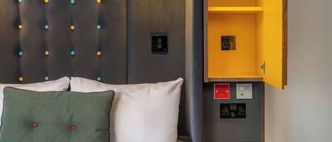 Una habitación de hotel con una cama, cabecera con panel de luz, mesita de noche con lámpara y unidad de almacenamiento integrada.