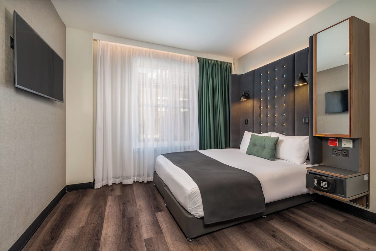 Hotellihuone, jossa on sänky, televisio, yöpöytä, peili ja ikkuna, jossa on verhot.