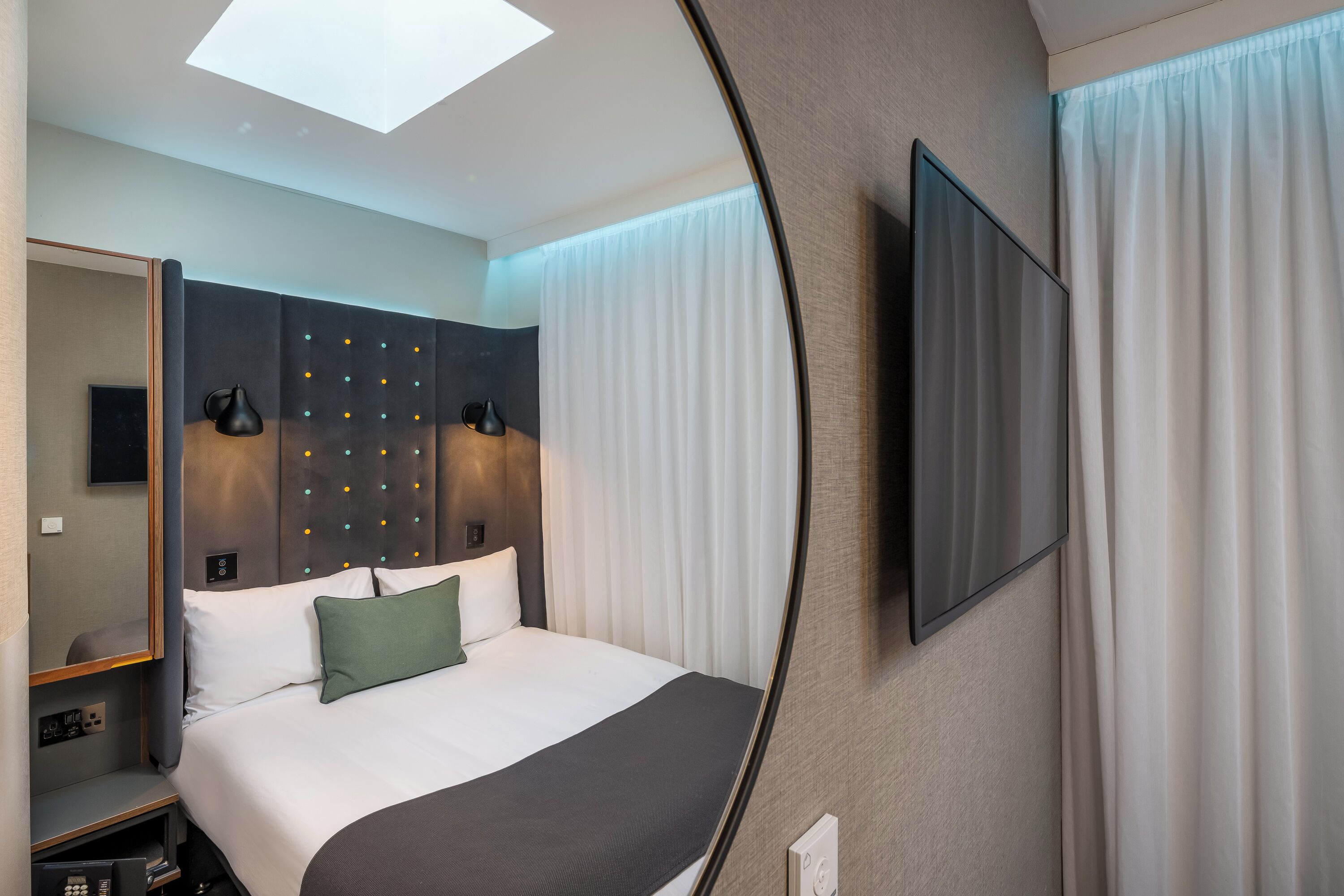A modern hotel room with a bed, a flat-screen TV, a mirror, and a ceiling light.