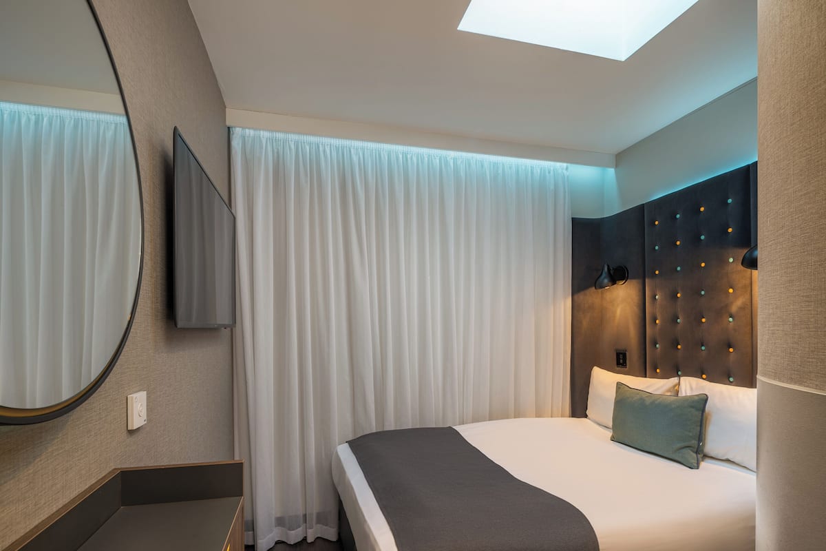 Hotellihuone, jossa on sänky, suuri peili, taulutelevisio ja kattovalaisin.