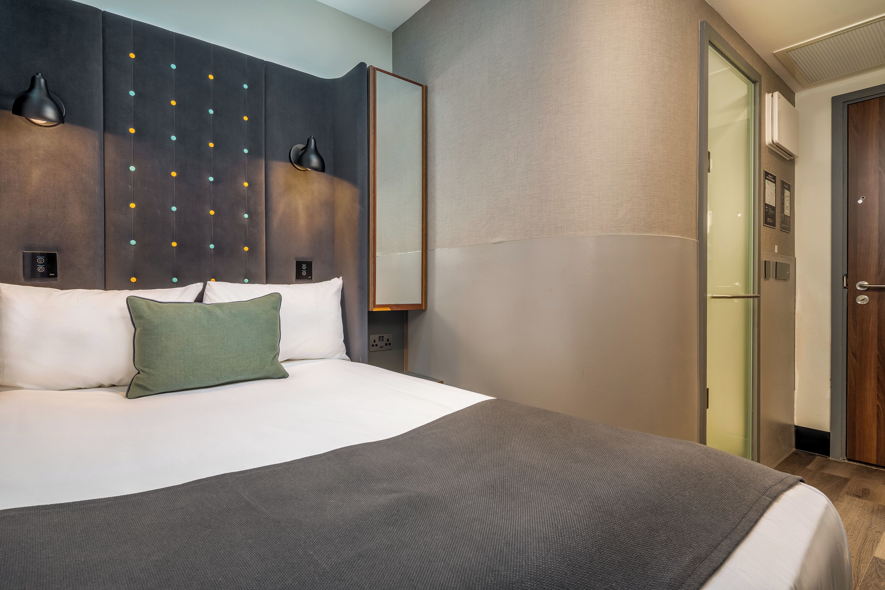 A hotel room with a large bed, a wall-mounted control panel, a mirror, and a door leading to a bathroom.