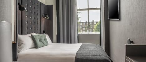 A modern hotel room with a large bed, a flat-screen TV, a bedside table, and a window with a view of buildings.