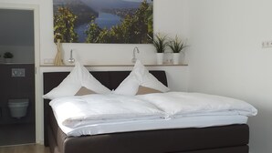 Uma cama bem arrumada com lençóis brancos, uma cabeceira escura e vista para um rio e montanhas pela janela.