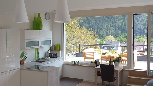 Cozinha moderna com armários brancos, sala de jantar com mesa e cadeiras, e varanda com vista para área arborizada.