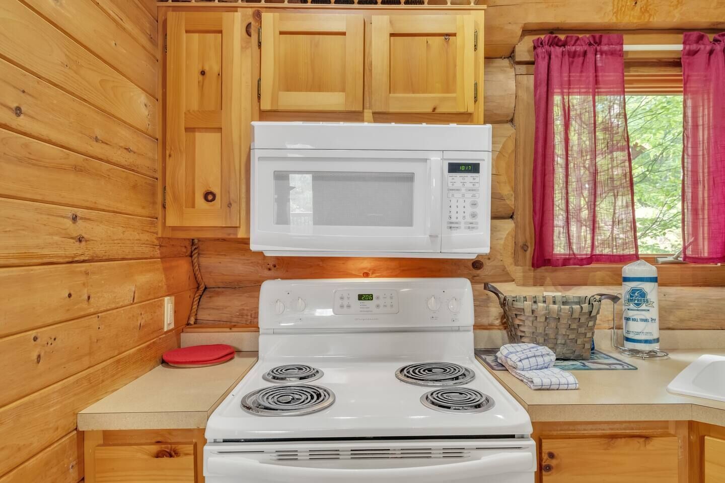 Una cocina compacta con paredes de madera, un microondas blanco, una estufa y una ventana con cortinas rosas.