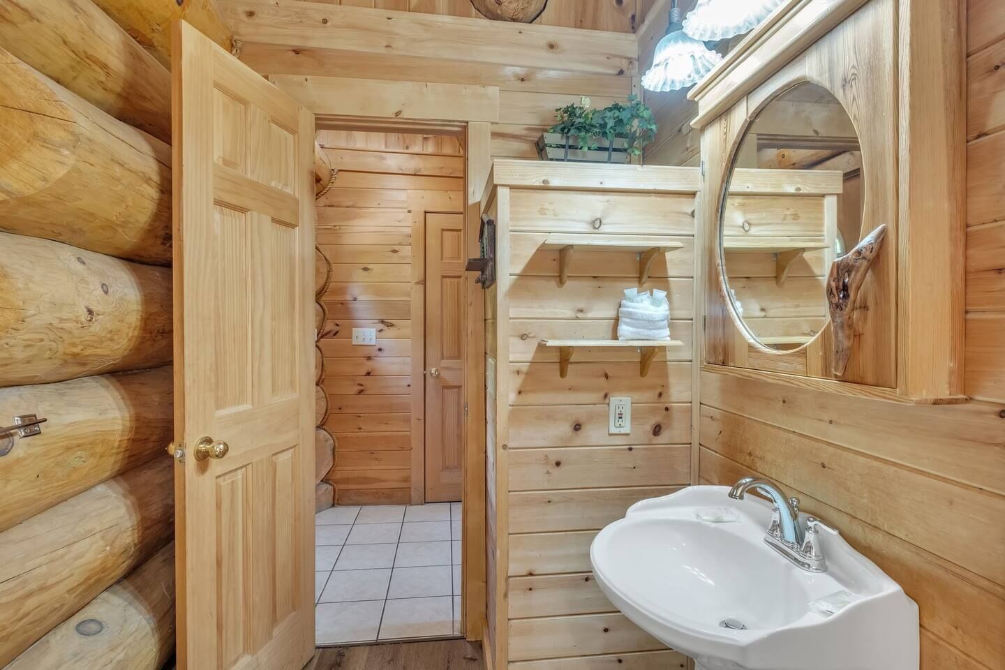 Baño de cabaña de madera con un lavamanos blanco, un espejo y una puerta que da a un pasillo con baldosas.