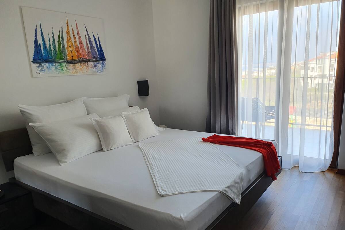 A neatly made bed with white linens and pillows, a red throw blanket, and a painting of sailboats on the wall.