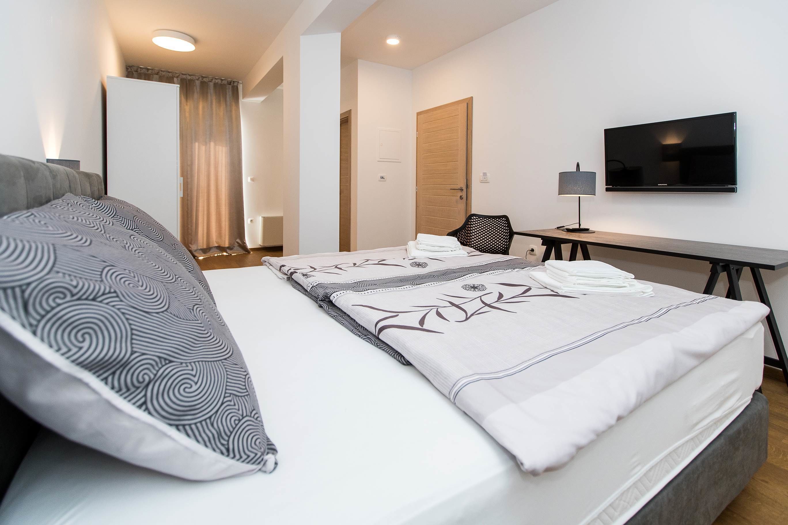 Una cama bien hecha con almohadas estampadas, un televisor de pantalla plana montado en la pared, un escritorio con lámpara y una silla.