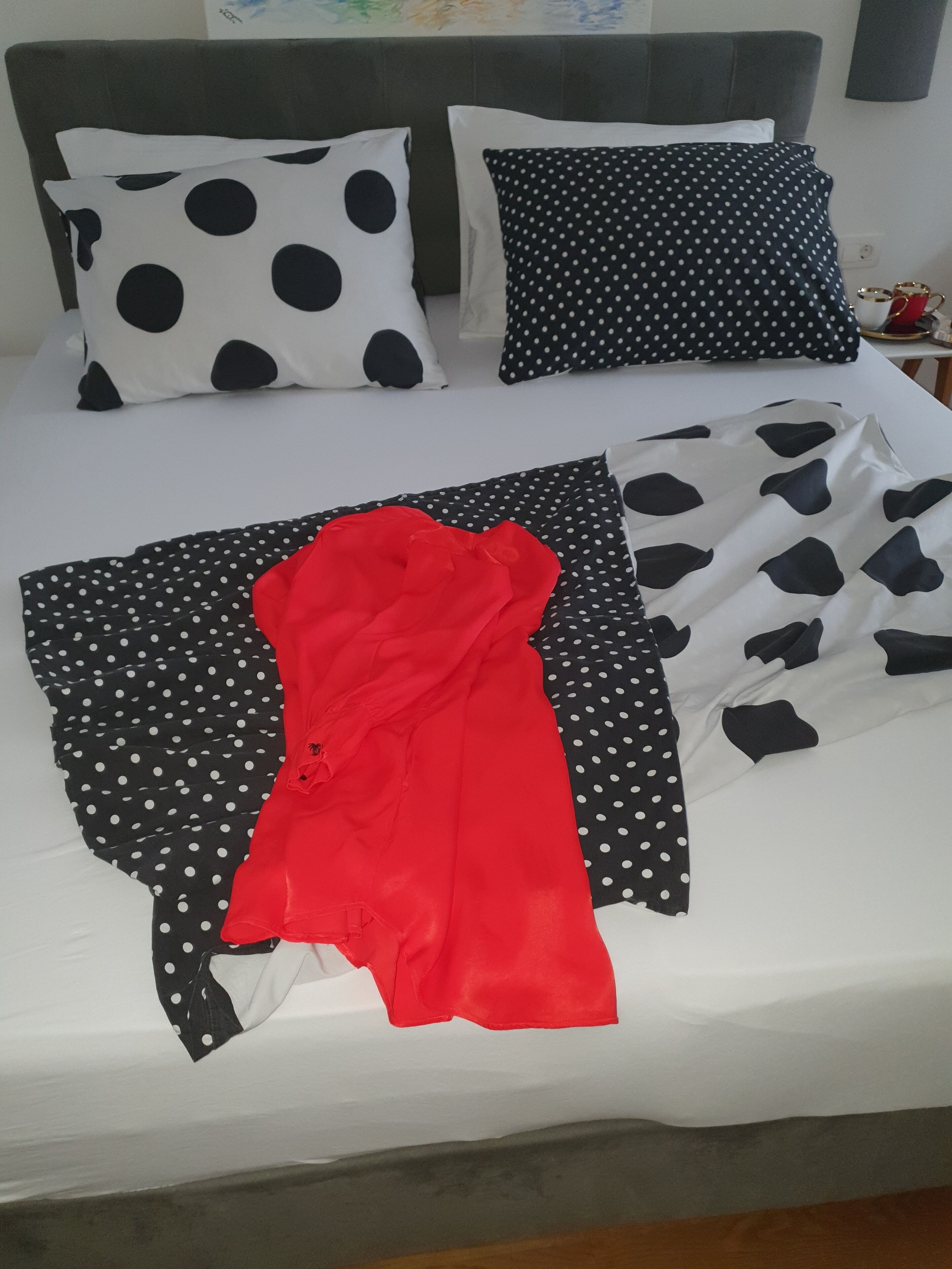 Een bed met zwart-wit stippenbeddengoed, een rode kledingstuk en een grijze hoofdbord.