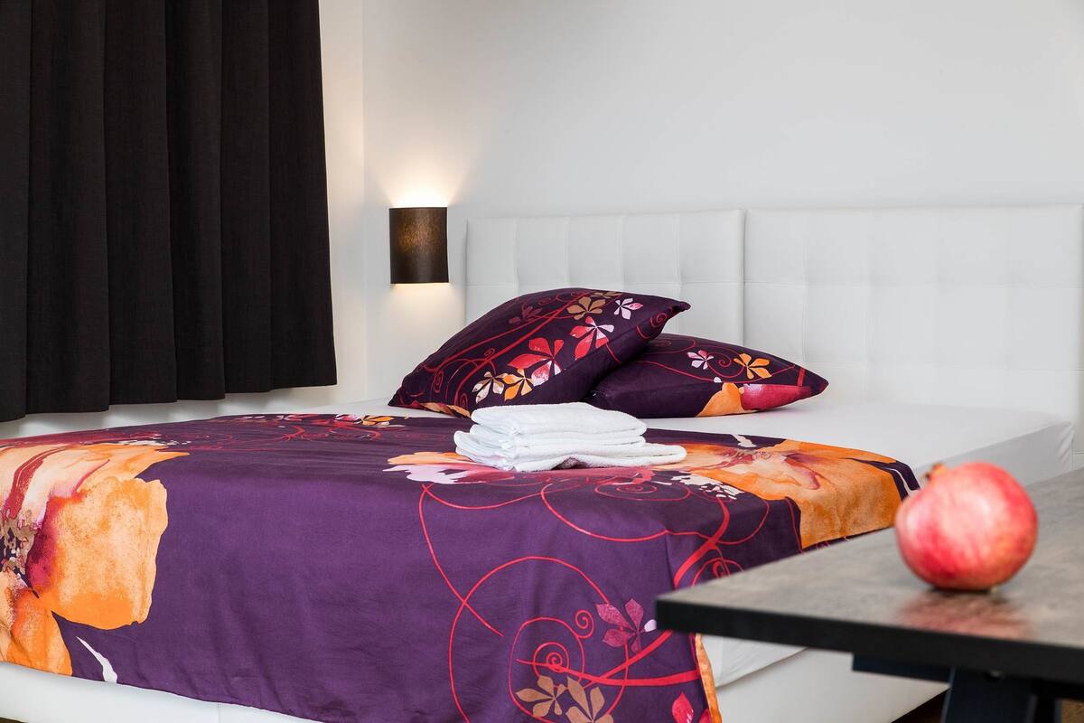 A neatly made bed with a purple floral bedspread, a white pillow, and a red apple on a table beside the bed.