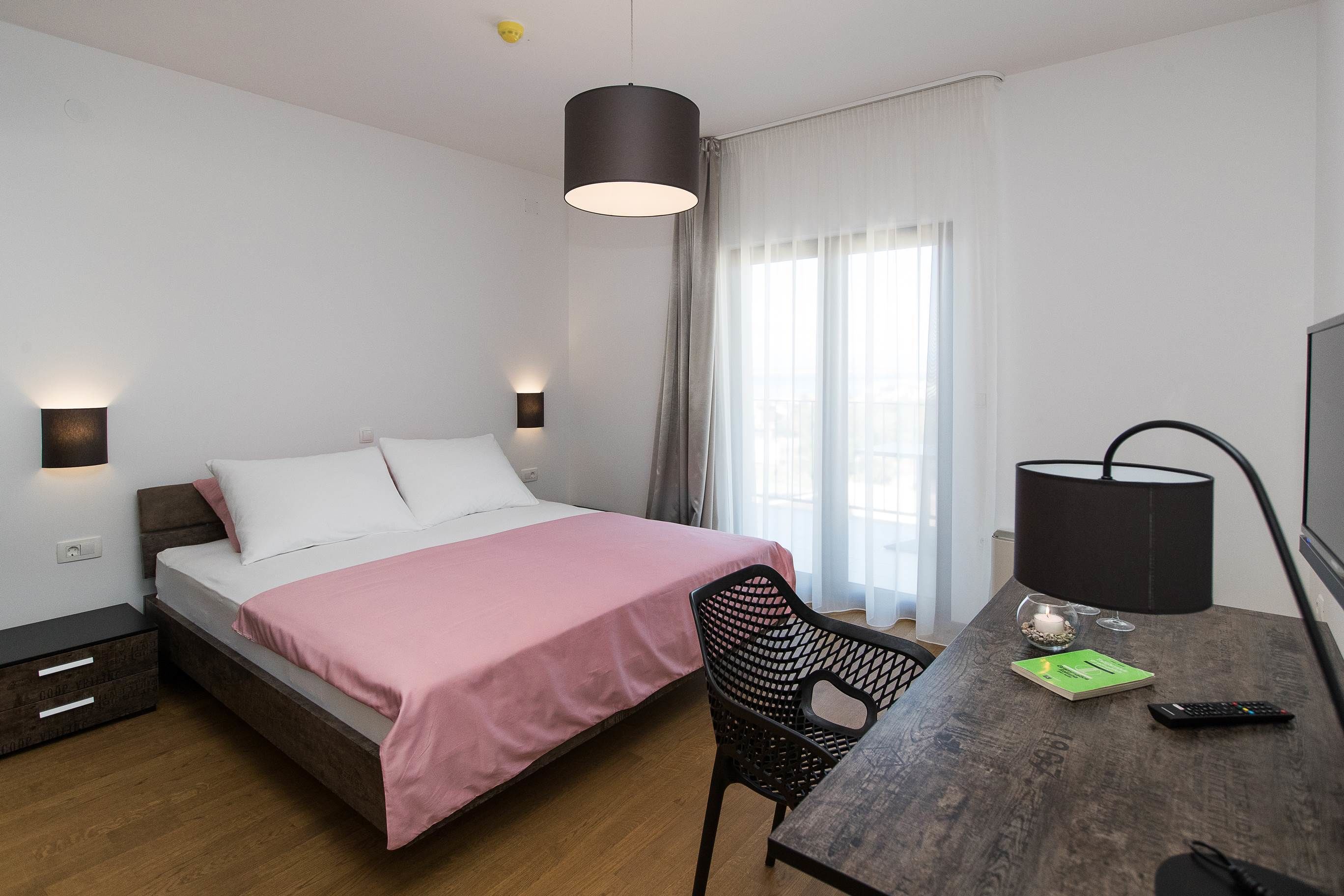 A bedroom with a bed, bedside tables, a desk with a chair, a lamp, and a window with curtains.