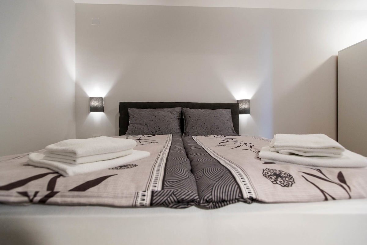 A neatly made bed with a patterned bedspread and pillows, two wall-mounted light fixtures, and a clean, minimalist room design.