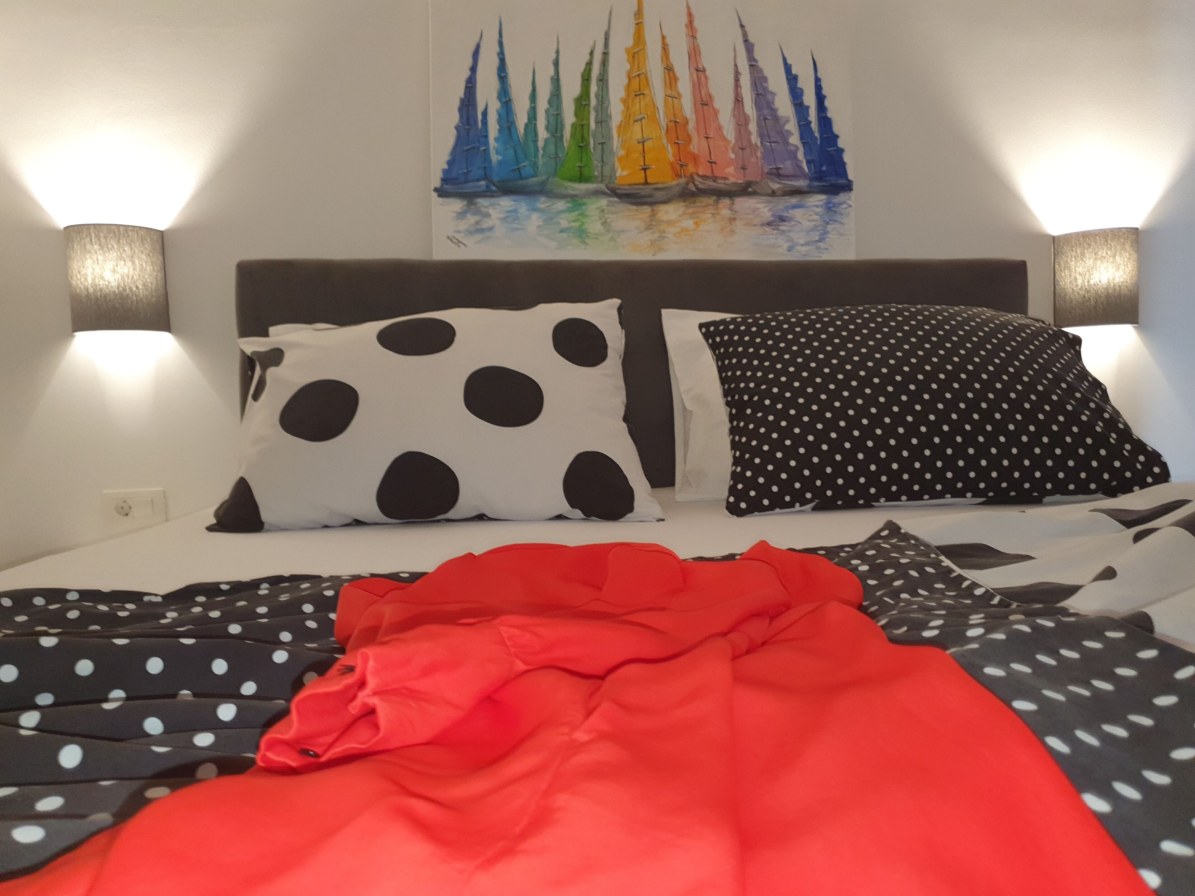 Una cama con almohadones a lunares y lisos, una colcha roja y un cuadro de un velero en la pared.